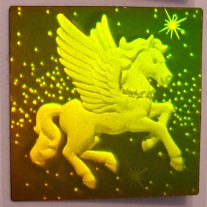 Puede incluir: Un Pegaso de color dorado con las alas extendidas, sobre un fondo verde oscuro con una dispersi&oacute;n de puntos blancos. El Pegaso est&aacute; en una pose de vuelo con la cabeza girada hacia un lado.