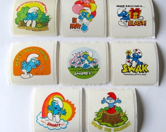 Puede incluir: Un conjunto de diez pegatinas vintage de los Pitufos. Las pegatinas presentan varios personajes y lemas de los Pitufos, incluyendo "Sé feliz", "Haz de cada día una explosión!" y "¡Todo está subiendo Pitufo!"