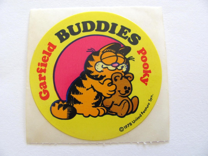 May include: A yellow circular sticker with the text "Garfield Buddies Pooky" in a black and red font. The sticker features a cartoon image of Garfield the cat hugging a brown teddy bear. The text "&copy;1978 United Feature Syn." is at the bottom of the sticker.