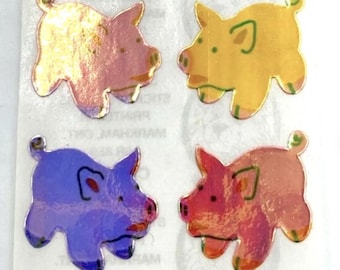 May include: Four individual pig-shaped stickers arranged in a grid on a white backing sheet. The stickers feature a metallic, iridescent finish in shades of pink, yellow, purple, and orange, each with simple line-drawn facial features.