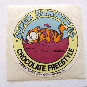 May include: A yellow and red circular sticker featuring Garfield the cat, a cartoon character, submerged in chocolate. The sticker reads "Super Summer '84 Chocolate Freestyle" and includes the copyright information "GARFIELD &copy; 1978 United Feature Syndicate, Inc."