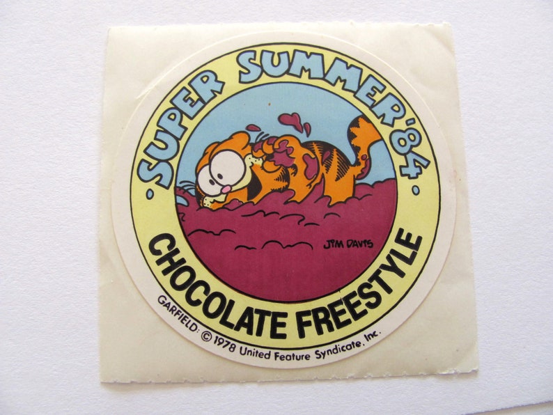 May include: A yellow and red circular sticker featuring Garfield the cat, a cartoon character, submerged in chocolate. The sticker reads "Super Summer '84 Chocolate Freestyle" and includes the copyright information "GARFIELD &copy; 1978 United Feature Syndicate, Inc."