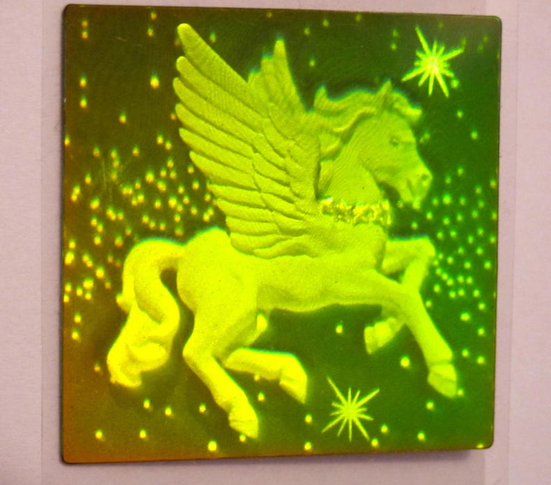 Puede incluir: Una pegatina hologr&aacute;fica verde que presenta un caballo alado con un collar dorado. El caballo se representa sobre un fondo verde oscuro con peque&ntilde;os puntos blancos.