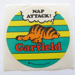 May include: A round Garfield sticker with a yellow, green, and white striped background. Garfield is sleeping and a thought bubble above his head says "Nap Attack!" The sticker has the copyright information "&copy;1978 United Feature Syn."