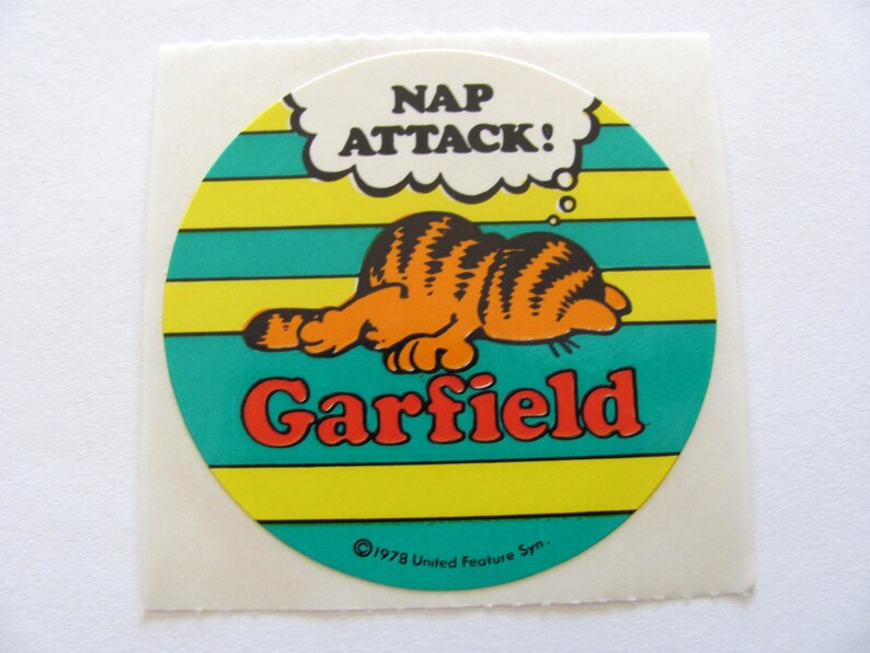May include: A round Garfield sticker with a yellow, green, and white striped background. Garfield is sleeping and a thought bubble above his head says "Nap Attack!" The sticker has the copyright information "&copy;1978 United Feature Syn."