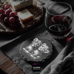 May include: A round, black slate coaster featuring a detailed illustration of two leopard cubs. The coaster is placed on a dark gray surface, with a glass of red wine and a basket of cheese and grapes in the background. The text "ILLUSION SlateStudio" is at the bottom.
