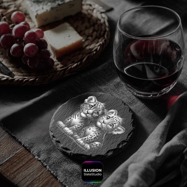 May include: A round, black slate coaster featuring a detailed illustration of two leopard cubs. The coaster is placed on a dark gray surface, with a glass of red wine and a basket of cheese and grapes in the background. The text "ILLUSION SlateStudio" is at the bottom.