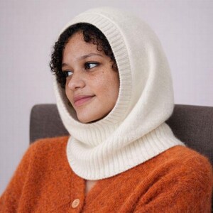 May include: Cream-colored knit balaclava. The hood covers the head and neck, with a ribbed design around the face and neck. The person in the image is wearing an orange cardigan. The balaclava is a winter accessory.