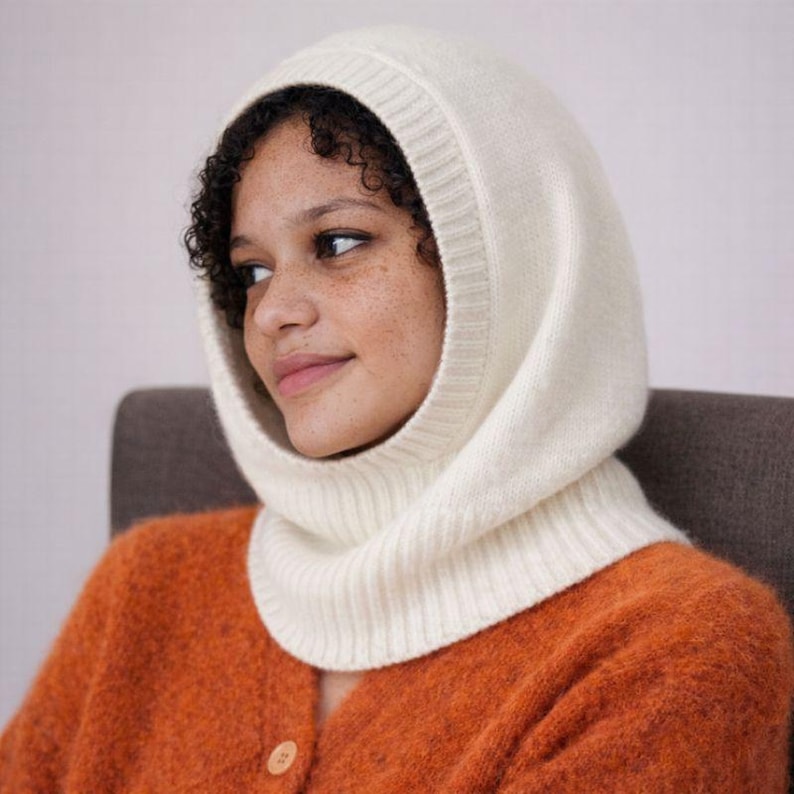 May include: Cream-colored knit balaclava. The hood covers the head and neck, with a ribbed design around the face and neck. The person in the image is wearing an orange cardigan. The balaclava is a winter accessory.