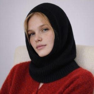 May include: A black knit balaclava, a type of head covering, is shown. The balaclava covers the head, neck, and part of the face. The person in the image is wearing a red sweater. The background is a neutral color.