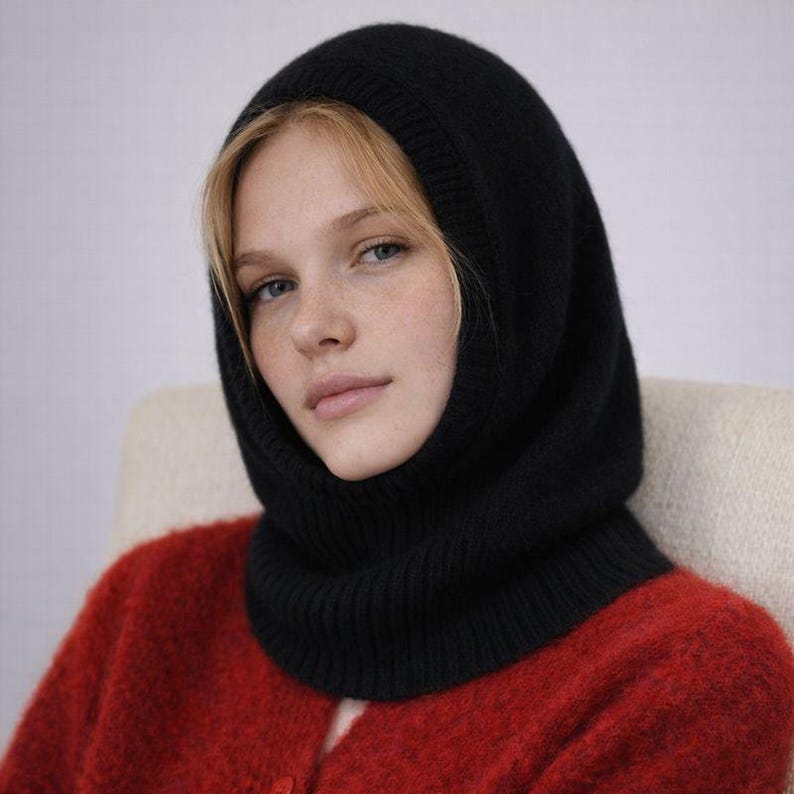May include: A black knit balaclava, a type of head covering, is shown. The balaclava covers the head, neck, and part of the face. The person in the image is wearing a red sweater. The background is a neutral color.