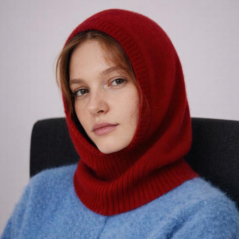 May include: A red knit balaclava, a type of head covering, is worn over a blue sweater. The balaclava covers the head and neck, leaving the face exposed. The knitwear has a ribbed texture around the neck and a smooth texture on the hood.