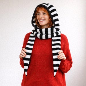 Może przedstawiać: Czarno-biały, pasiasty zestaw dzianinowy z kapturem i szalikiem. Kaptur jest przymocowany do szalika, tworząc spójny design. Szalik zwisa z przodu. Zestaw wykonany jest z miękkiego, teksturowanego materiału.