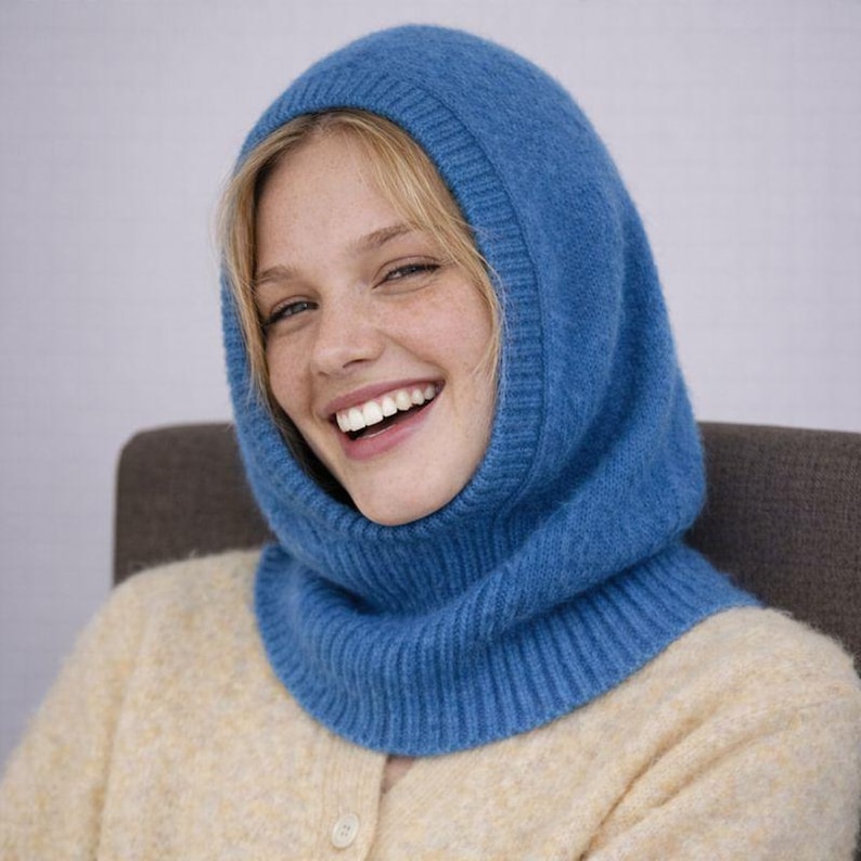 May include: A blue knitted balaclava with a ribbed neck and hood. The balaclava is made of a soft, textured material. The person in the image is wearing a light yellow cardigan.