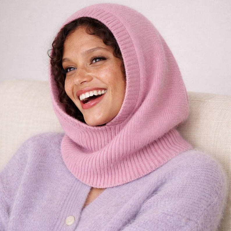 May include: A light pink knitted balaclava-style hood. The hood is made of a soft, ribbed knit material. The person in the image is wearing a light purple cardigan. The hood covers the head and neck, leaving the face exposed.