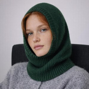 May include: A dark green knit balaclava. The balaclava covers the head and neck, with an opening for the face. The knitwear is a solid, deep green color. The person in the image is wearing a gray sweater.