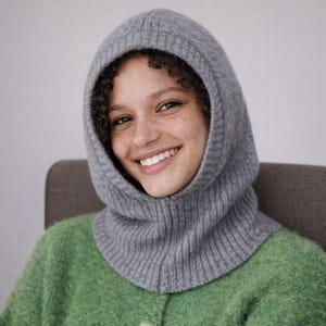 May include: A gray knitted balaclava with a ribbed texture. The hood covers the head and neck, with an opening for the face. The balaclava is made of a soft, warm material, perfect for cold weather. The person in the image is wearing a green sweater.