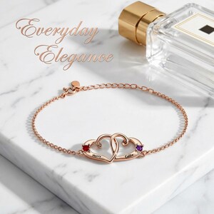 May include: Rose gold bracelet featuring two intertwined heart-shaped pendants, each adorned with a gemstone. The bracelet includes the names "Herry" and "Callie". The words "Everyday Elegance" are displayed in the upper left corner.