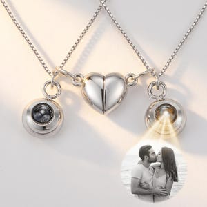 Puede incluir: Un conjunto de collares en tono plateado con un colgante central en forma de corazón que se divide en dos. Cada mitad está unida a un dije circular con una lente de proyección. Una lente proyecta una fotografía en blanco y negro de una pareja.