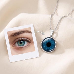 Op de afbeelding: Een zilverkleurige ketting met een ronde hanger met een realistische blauwe iris en zwarte pupil. De hanger zit aan een fijne zilveren ketting. Naast de ketting ligt een vierkante foto van een menselijk oog.