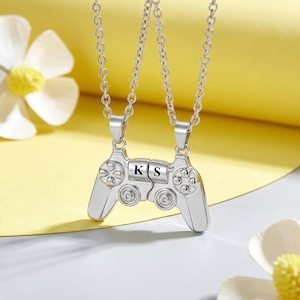 May include: Two silver-colored pendant necklaces. Each pendant is shaped like a video game controller. The controllers are connected, with the letters "K" and "S" engraved on them. The necklaces hang on a silver chain.