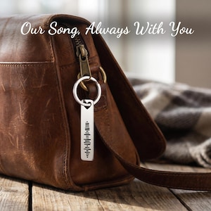 May include: A silver keychain with a barcode design hangs from a brown leather purse. The keychain is rectangular with a series of vertical lines. The purse has a zipper and a strap. The text "Our Song, Always With You" is at the top.