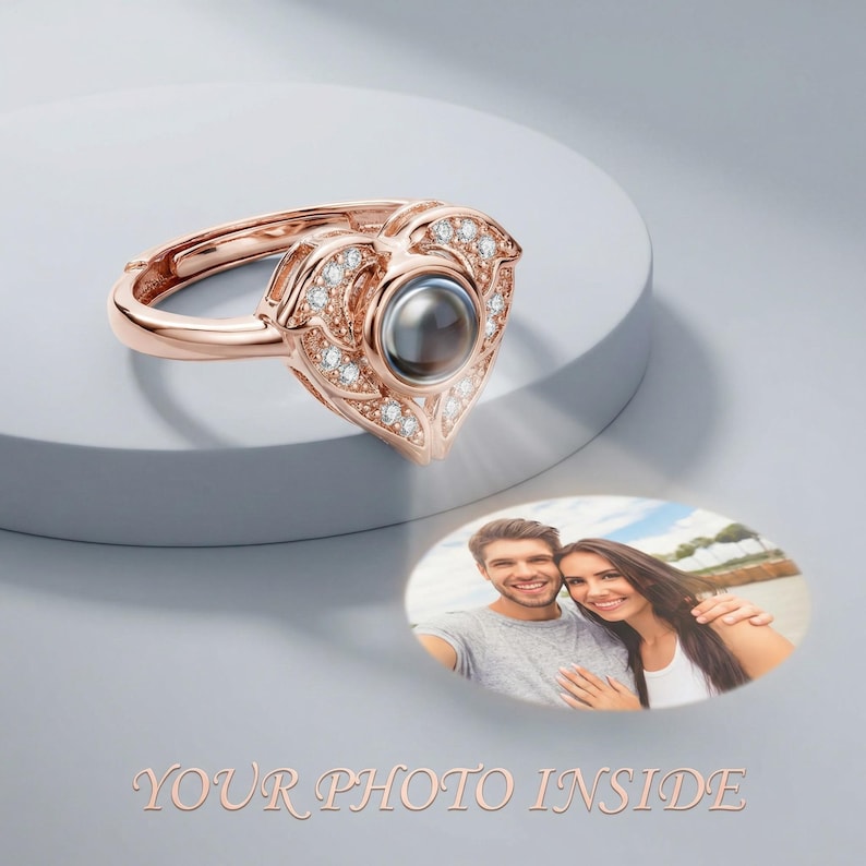 Puede incluir: Anillo en forma de coraz&oacute;n de oro rosa con piedras preciosas transparentes y una piedra oscura central. El anillo proyecta una foto de una pareja. El texto en la parte inferior dice "YOUR PHOTO INSIDE".