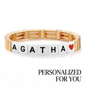 May include: A stretch bracelet featuring gold-toned rectangular links and white square beads spelling the name AGATHA in black capital letters, finished with a small red heart icon. The text PERSONALIZED FOR YOU appears below the bracelet.