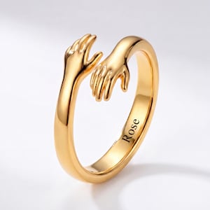 May include: A gold-toned open ring featuring two hands reaching towards each other in a hugging design. The inner band of the ring is engraved with the name Rose in a clear, black, sans-serif font, creating a smooth and polished metallic finish.