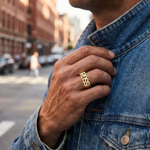 May include: A gold-colored ring with the name "ROSS" in block letters. The ring is worn on a finger, set against a denim jacket background. The ring is a personalized jewelry item.