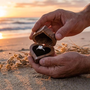 Può includere: Un portagioie in legno a forma di cuore, aperto per rivelare un anello di fidanzamento con diamanti. La scatola è tenuta in mano su uno sfondo di spiaggia al tramonto. L'anello presenta un diamante a taglio quadrato.