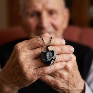 May include: A black heart-shaped pendant necklace with a black chain. The pendant features a black and white photograph of a person. The necklace is held in a person's hands.