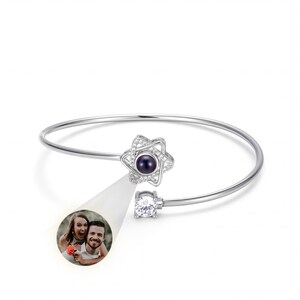 Può includere: Un braccialetto a cerchio color argento con un ciondolo a forma di stella con una pietra centrale viola scuro e cristalli trasparenti. Una gemma rotonda e trasparente è incastonata all'estremità opposta. Viene proiettata una piccola immagine di una coppia.