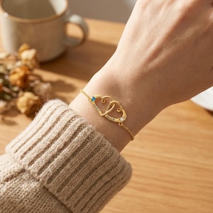 May include: A gold bracelet with two interlocking heart pendants. Each heart is inscribed with a name and set with a gemstone. The bracelet is attached by a fine chain, creating a personalised and elegant accessory.