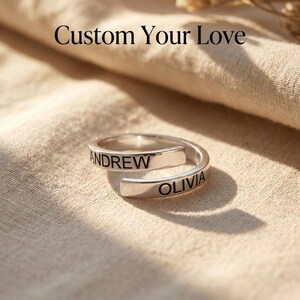 Może przedstawiać: Srebrny pierścionek typu wrap z wygrawerowanymi imionami "ANDREW" i "OLIVIA". Pierścionek jest prezentowany na neutralnej tkaninie, z tekstem "Custom Your Love" powyżej. Spersonalizowana biżuteria.