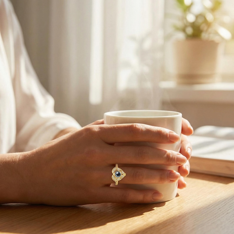 Puede incluir: Un anillo de oro con un dise&ntilde;o en forma de coraz&oacute;n, con una piedra central azul oscuro y piedras m&aacute;s peque&ntilde;as alrededor. El anillo se lleva en un dedo, con una taza blanca en las manos. La taza humea, sugiriendo una bebida caliente.