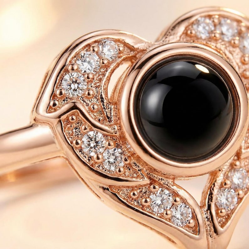Puede incluir: Anillo de oro rosa con dise&ntilde;o de coraz&oacute;n. El anillo presenta una piedra negra grande y redonda en el centro, rodeada de piedras m&aacute;s peque&ntilde;as, claras y brillantes. La banda y el engaste tienen un intrincado patr&oacute;n en forma de hoja.