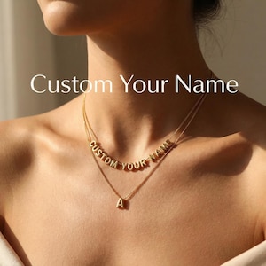 以下が含まれることがあります： 画像には「Custom Your Name」の文字が入ったゴールドカラーのネックレス。 ネックレスは2層になっており、上層はフレーズを綴り、下層は1文字のペンダントが付いています。 ネックレスは人の首にかけられています。