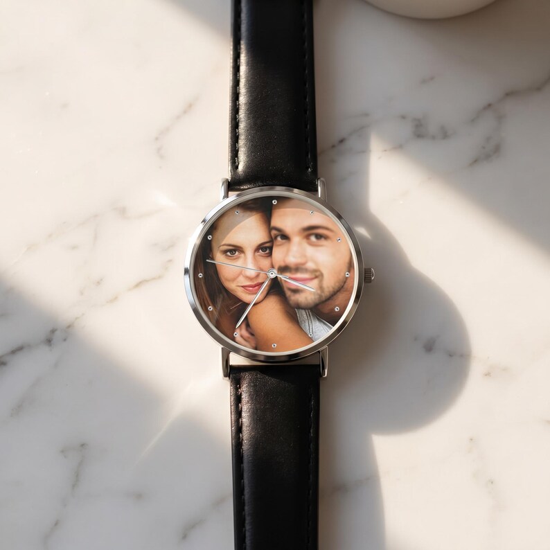 Peut inclure: Une montre-bracelet avec un bracelet en cuir noir et un bo&icirc;tier circulaire argent&eacute;. Le cadran de la montre pr&eacute;sente une photographie couleur personnalis&eacute;e d'un couple, avec de petits marqueurs argent&eacute;s et de fines aiguilles argent&eacute;es.