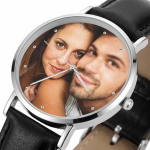 Peut inclure: Une montre analogique ronde avec un bo&icirc;tier en m&eacute;tal argent&eacute; et un bracelet en cuir noir. Le cadran de la montre pr&eacute;sente une impression photographique personnalis&eacute;e d'un couple, agr&eacute;ment&eacute;e de petits index argent&eacute;s et d'aiguilles fines.