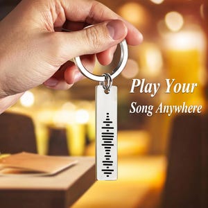 May include: A silver keychain with a rectangular tag featuring a sound wave design. The text "Play Your Song Anywhere" is visible in the background. The keychain is held up against a blurred background.