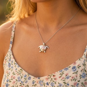 Puede incluir: Un collar de plata con un colgante de tortuga. El colgante presenta un diseño detallado de tortuga con un caparazón rosa y dorado. El collar lo lleva una persona que lleva una blusa con estampado floral.