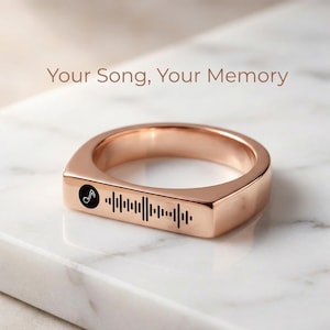 May include: Rose gold ring with a rectangular design. The ring features a black musical note icon and a barcode-like pattern. The text "Your Song, Your Memory" is displayed above the ring.
