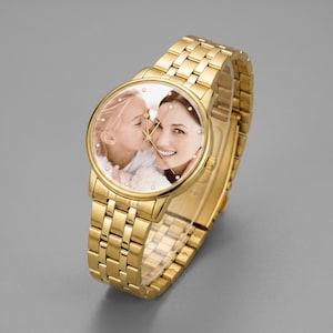 May include: Gold-toned wristwatch with a photo of a woman and a child on the watch face. The watch has a gold band and a round face with small, decorative crystals. The watch is displayed on a gray background.