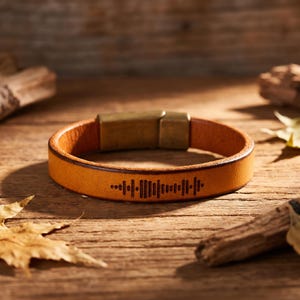 May include: A tan leather bracelet with a gold-colored clasp. The bracelet features a laser-etched sound wave design. The bracelet is displayed on a wooden surface with leaves and wood pieces.