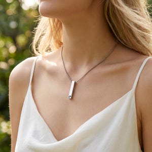 May include: A silver bar pendant necklace with a small, round accent stone. The pendant hangs from a delicate ball chain. The necklace is worn against a white top, with a blurred green background.