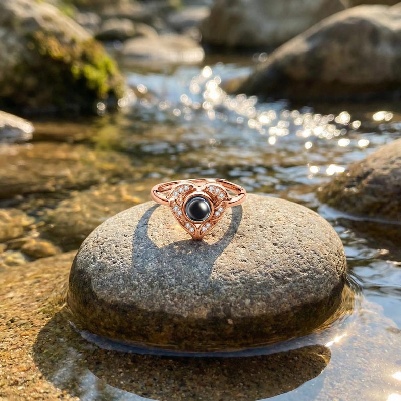 Puede incluir: Anillo de oro rosa con dise&ntilde;o de coraz&oacute;n. El anillo presenta una piedra central negra rodeada de peque&ntilde;as piedras preciosas transparentes. El anillo est&aacute; colocado sobre una piedra gris, con agua y rocas en el fondo.