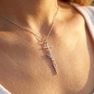May include: A silver necklace with a cross-shaped pendant. The pendant has the words "my person" written vertically. The necklace is suspended from a fine chain. The background is blurred.