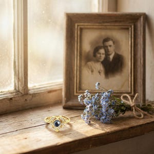 Puede incluir: Un anillo dorado con dise&ntilde;o de coraz&oacute;n, con una piedra central azul oscuro y piedras claras circundantes. El anillo est&aacute; sobre una superficie de madera junto a una fotograf&iacute;a vintage enmarcada y un peque&ntilde;o ramo de flores azules.