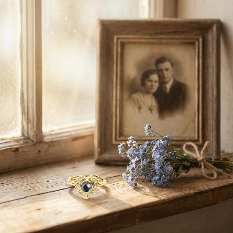 Puede incluir: Un anillo dorado con dise&ntilde;o de coraz&oacute;n, con una piedra central azul oscuro y piedras claras circundantes. El anillo est&aacute; sobre una superficie de madera junto a una fotograf&iacute;a vintage enmarcada y un peque&ntilde;o ramo de flores azules.